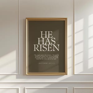 Op de afbeelding: Een ingelijste print met de woorden "HE HAS RISEN" in witte letters op een donker olijfgroene achtergrond. De print bevat een citaat uit Mattheüs 28:5-6. De lijst is goudkleurig.