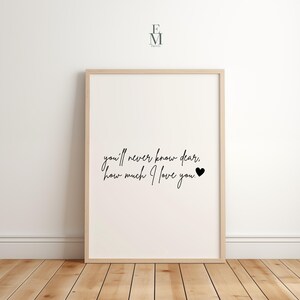 May include: A framed print with the text "you'll never know dear, how much I love you." in black script. The print is in a light wood frame and is displayed against a white wall and wooden floor.
