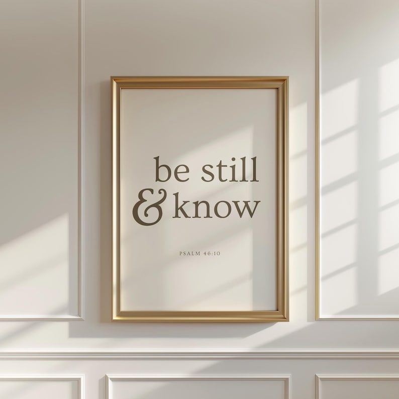 May include: A framed print with the words "be still & know" in a stylish font. The text is in a dark brown color against a cream background. The frame is gold, and the print is mounted on a white wall.