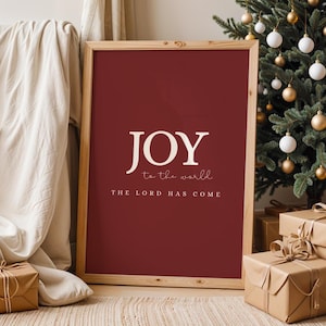 May include: A framed print with a burgundy background and the words "JOY to the world THE LORD HAS COME" in white. The print is in a wooden frame and is displayed near a Christmas tree and wrapped gifts.