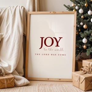 Könnte beinhalten: Ein gerahmter Druck mit dem Wort "JOY" in burgunderfarbener Schrift, darunter "to the world" und "THE LORD HAS COME". Der Druck befindet sich in einem hellen Holzrahmen, mit einem Weihnachtsbaum und verpackten Geschenken im Hintergrund.
