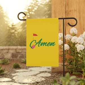May include: A yellow garden flag with the word "Amen" in teal script, a red flag, and a pink flower graphic. The flag is displayed on a black metal stand in a garden setting.