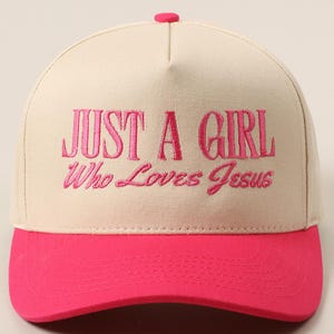 May include: A baseball cap with a cream-colored crown and a bright pink brim. The cap features the embroidered text "JUST A GIRL Who Loves Jesus" in pink thread. The top button is also pink.