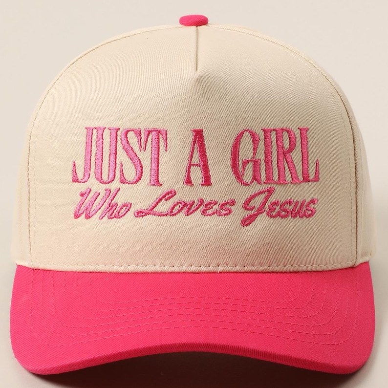 May include: A baseball cap with a cream-colored crown and a bright pink brim. The cap features the embroidered text "JUST A GIRL Who Loves Jesus" in pink thread. The top button is also pink.