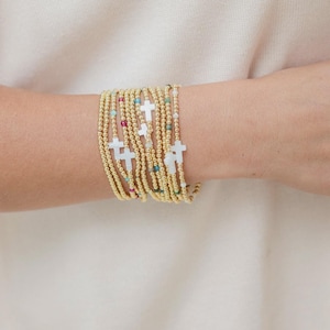 May include: A stack of multiple gold-toned beaded bracelets featuring small white cross charms and interspersed colorful accent beads. The bracelets are worn together, creating a layered appearance with metallic gold spheres and white crosses.