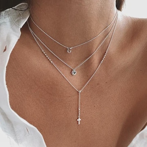 May include: A layered silver necklace set consisting of three delicate chains. The top chain features a small letter G pendant, the middle chain has a tiny heart charm, and the bottom chain is a lariat style with a small cross pendant at the end.