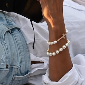 May include: Two pearl bracelets adorn a wrist, one featuring a gold cross pendant. The other bracelet is made of round pearls. The person is wearing a white shirt and holding a black book with the text "NKJV".