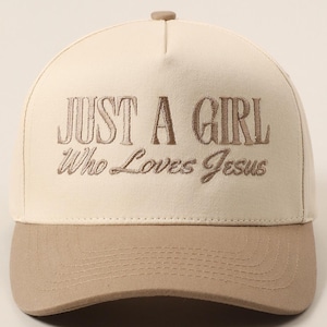 May include: A beige baseball cap with a curved brim and a button on top. The cap features the embroidered text "JUST A GIRL Who Loves Jesus" in a brown color.