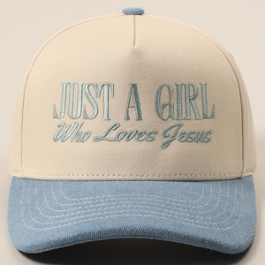 May include: A baseball cap with a light beige crown and a light blue brim. The cap features the embroidered text "JUST A GIRL Who Loves Jesus" in a matching light blue color. The cap has a small blue button on top.