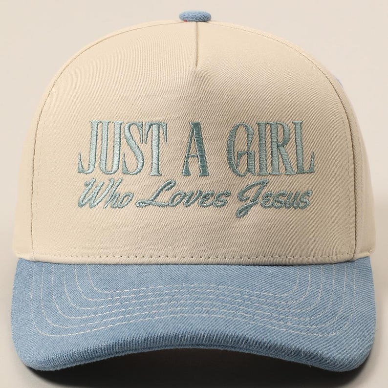 May include: A baseball cap with a light beige crown and a light blue brim. The cap features the embroidered text "JUST A GIRL Who Loves Jesus" in a matching light blue color. The cap has a small blue button on top.