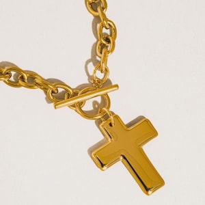 May include: A gold-colored necklace with a cross pendant. The necklace features a chunky chain design with a toggle clasp. The cross pendant is a solid, polished gold color. The jewelry is displayed on a white background.