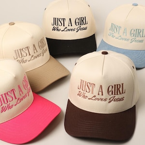 May include: Cream-colored baseball caps with the embroidered text "JUST A GIRL Who Loves Jesus." The caps have different colored brims, including black, brown, pink, tan, and light blue. The caps are arranged on a light surface.