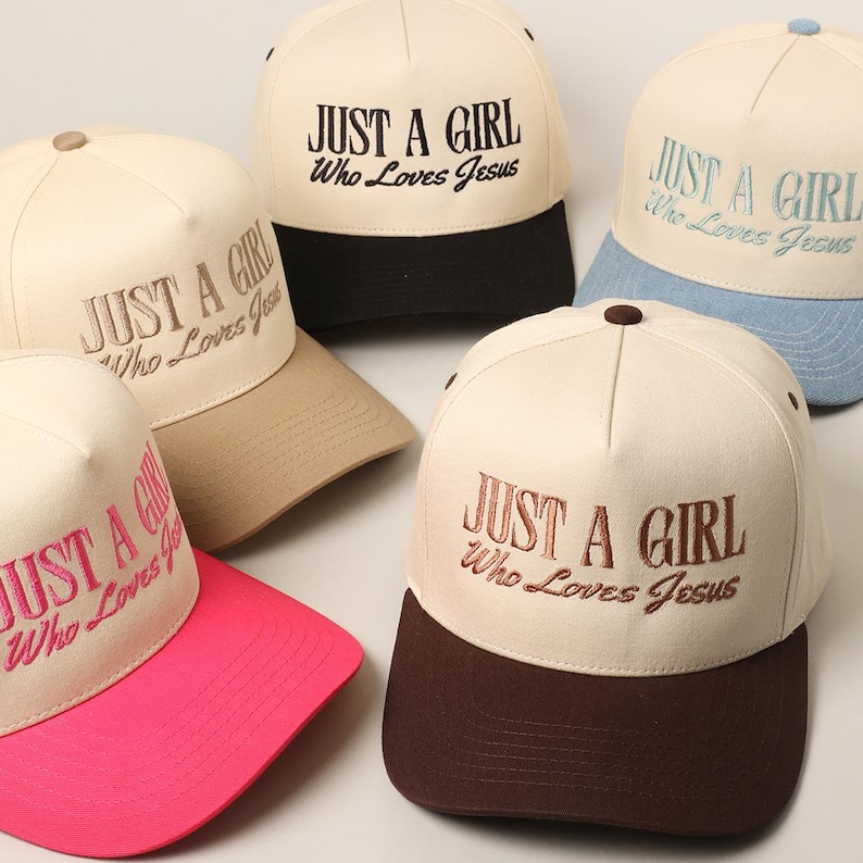 May include: Cream-colored baseball caps with the embroidered text "JUST A GIRL Who Loves Jesus." The caps have different colored brims, including black, brown, pink, tan, and light blue. The caps are arranged on a light surface.