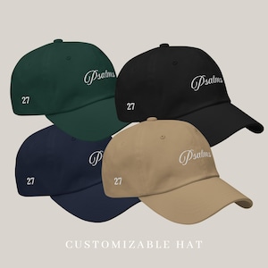 以下が含まれることがあります： ダークグリーン、ブラック、ネイビーブルー、タンの4つの野球帽。各帽子には、前面に白い筆記体で「Psalms」の文字が刺繍され、側面に数字の「27」が刺繍されています。「CUSTOMIZABLE HAT」というテキストが下にあります。