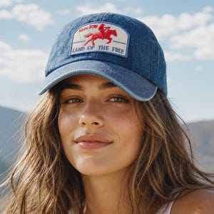 May include: A blue denim baseball cap with a curved brim. The cap features a rectangular patch with a red and white embroidered design of a horse and rider, along with the text "Since 1776" and "Land of the Free".