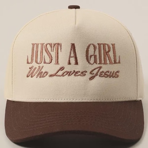 May include: A baseball cap with a cream-colored crown and a brown brim. The cap features the embroidered text "JUST A GIRL Who Loves Jesus" in a brown color. The top button is also brown.