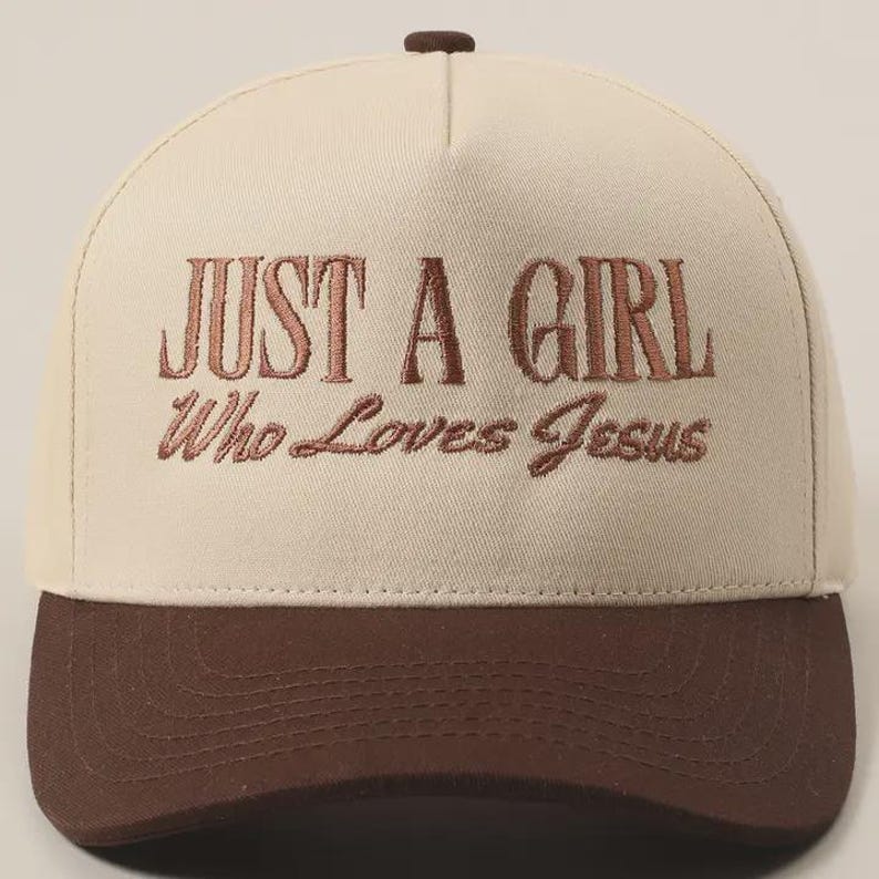 May include: A baseball cap with a cream-colored crown and a brown brim. The cap features the embroidered text "JUST A GIRL Who Loves Jesus" in a brown color. The top button is also brown.