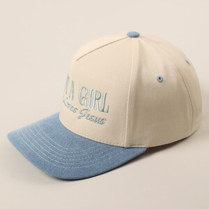 May include: A baseball cap with a cream-colored crown and a light blue brim. The cap features the embroidered text "A GIRL Loves Jesus" in light blue thread. The cap has a button on top and a curved brim.
