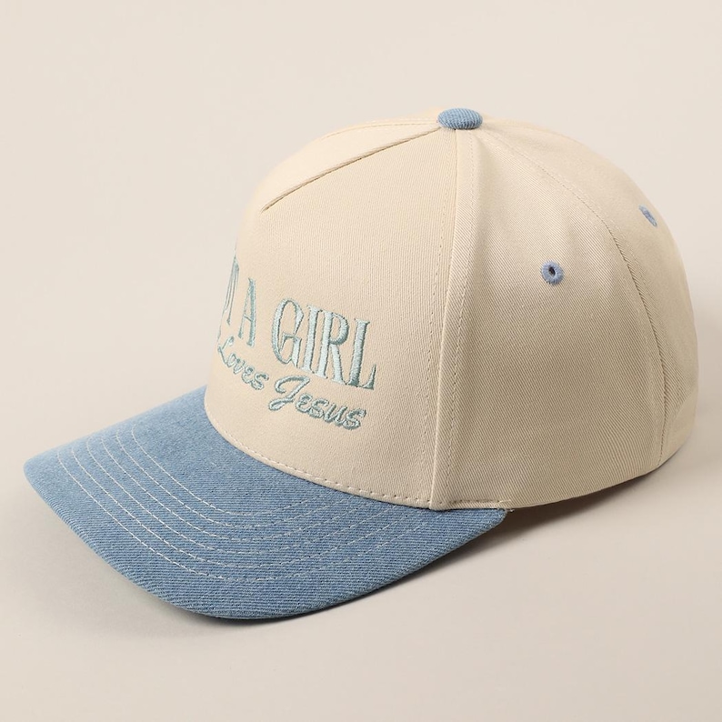 May include: A baseball cap with a cream-colored crown and a light blue brim. The cap features the embroidered text "A GIRL Loves Jesus" in light blue thread. The cap has a button on top and a curved brim.