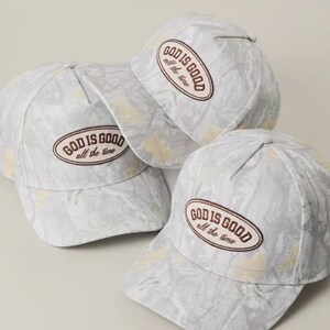 May include: Several white baseball caps with a subtle floral pattern. Each cap features the embroidered text "GOD IS GOOD all the time" in a brown oval. The caps are arranged in a casual, overlapping manner.