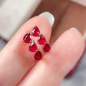 May include: A cluster of six teardrop-shaped, deep red gemstones set in gold. The gems are arranged in a triangular pattern, showcasing their vibrant color and brilliance. The jewelry is held in a person's hand.