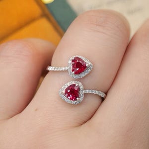 May include: A bypass ring featuring two heart-shaped red gemstones, each surrounded by a halo of small white stones. The band is made of a silver-toned metal and is encrusted with tiny white stones along the visible portion of the shank.