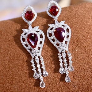 May include: Dangle earrings with large, pear-shaped, deep red gemstones. The earrings have a diamond-like, clear, sparkling, encrusted design. The earrings have a diamond-like, clear, sparkling, encrusted design with dangling teardrop-shaped accents.