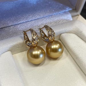 May include: A pair of gold-colored earrings with a large, round, golden pearl pendant. The earrings have a decorative gold clasp with small, clear gemstones. The earrings are displayed in a white jewelry box.