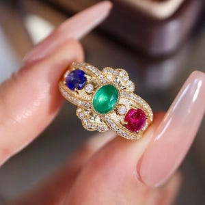 Könnte beinhalten: Ein goldener Ring mit einem ovalen grünen Edelstein in der Mitte, flankiert von einem blauen und einem roten Edelstein. Der Ring ist mit kleinen, funkelnden Diamanten besetzt, die ein luxuriöses Design schaffen. Der Ring wird von einer Hand gehalten.