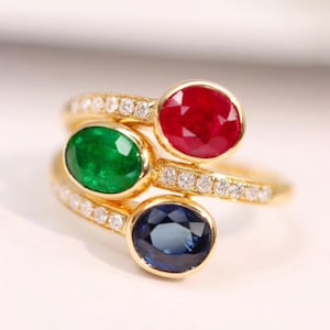 May include: A gold ring with three oval gemstones: a red ruby, a green emerald, and a blue sapphire. The ring band is adorned with small, sparkling diamonds, creating a luxurious and elegant design.
