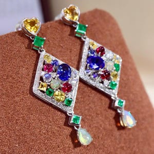 Pode incluir: Um par de brincos pendurados deslumbrantes. Cada brinco apresenta uma pedra preciosa amarela em forma de coração no topo, uma pedra preciosa verde quadrada e um pingente em forma de diamante. Os pingentes são adornados com uma variedade de pedras preciosas coloridas, incluindo pedras azuis, vermelhas e amarelas, e uma opala em forma de gota.