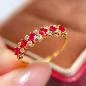 May include: A gold ring featuring a row of square-cut red gemstones alternating with small, round, clear gemstones. The ring has a delicate, elegant design, with a polished gold band.