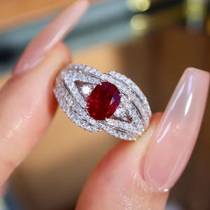 Può includere: Un anello sbalorditivo con un grande rubino ovale al centro, circondato da un alone di gemme chiare e scintillanti. La fascia dell'anello è finemente progettata con più file di pietre più piccole, creando un gioiello lussuoso e accattivante.