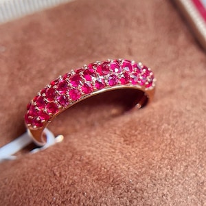 May include: A gold ring with a band covered in small, vibrant red gemstones. The ring is set against a brown background, highlighting the color and sparkle of the stones. The gemstones are closely set, creating a continuous surface of red.