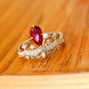 Könnte beinhalten: Ein silberner Ring mit einem birnenförmigen, tiefroten Edelstein. Der Ring hat ein kronenartiges Design mit kleinen, klaren Edelsteinen und dekorativen Elementen. Der Ring liegt auf einer Holzoberfläche.