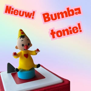 Op de afbeelding: Een gele speelgoedclown met een rode neus en een rood hart op zijn borst zit op een blauwe basis. De clown houdt rode ballen in elke hand. Het speelgoed staat op een rode en witte doos. De tekst "Nieuw! Bumba tonie!" staat in rood en roze op een regenboogachtergrond.