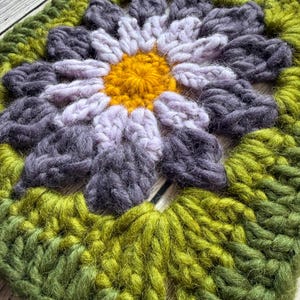 May include: A square, handmade crochet piece featuring a large flower design. The flower has a yellow center, surrounded by petals in shades of lavender and gray. The outer border is green, creating a vibrant, textured textile.
