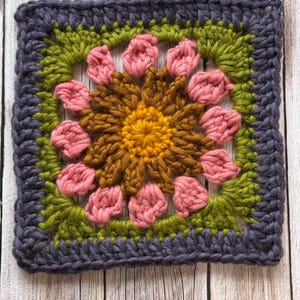 May include: A square, handmade crochet piece featuring a floral design. The center is yellow, surrounded by brown petals, pink flower petals, green leaves, and a dark gray border. The piece is likely part of a larger craft project.