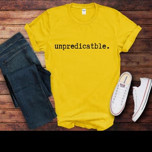 May include: A yellow t-shirt with the text "unpredictable." printed in black on the front. The shirt is laid flat on a wooden surface with a pair of blue jeans and white sneakers.