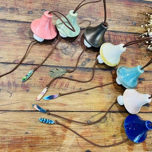 May include: A selection of ceramic bottle stoppers in shades of pink, green, black, yellow, blue, and white. Each stopper has a leather cord and a decorative charm. The charms include a leaf and patterned pendants. The stoppers are designed for wine bottles.