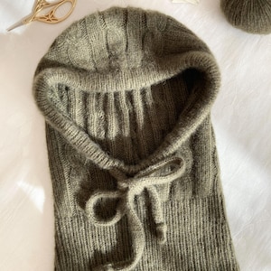 May include: A knitted olive green balaclava with a drawstring closure. The balaclava is made of a soft, textured yarn and features a ribbed knit design. A pair of gold scissors and a ball of yarn are in the background.
