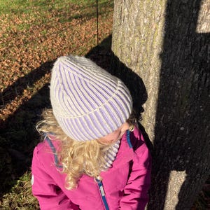 Könnte beinhalten: Eine hellgrau-lila gestreifte Strickmütze und ein passender Schal. Die Mütze ist gerippt und hat einen umgeschlagenen Rand. Die Person trägt eine leuchtend pinkfarbene Jacke mit blauen Handschuhen. Der Hintergrund umfasst einen Baumstamm und Gras.