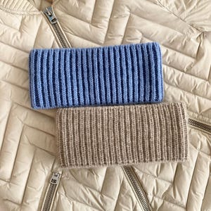 May include: Two ribbed knit headbands, one light blue and one beige, are displayed on a cream-colored quilted jacket. The headbands are rectangular and made of a thick, textured yarn. The jacket has a zipper closure.
