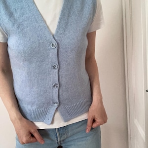 Puede incluir: Un chaleco de punto azul claro con botones y cuello en V, usado sobre una camiseta blanca y vaqueros azules claros. El chaleco tiene un dobladillo acanalado y está hecho de un material de punto suave. Se cierra con botones redondos.
