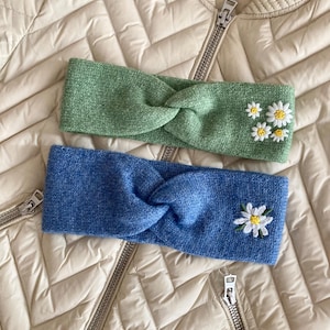May include: Two knitted headbands with a front twist detail. The top headband is sage green with four embroidered white daisy flowers. The bottom headband is cornflower blue with a single embroidered white daisy flower with a yellow center.