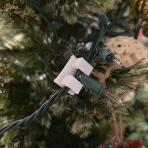Puede incluir: Primer plano de un árbol de Navidad con agujas verdes y clips de luz de plástico blanco. Los clips sujetan las luces navideñas con cable verde en su lugar. Un adorno de coche rojo y un adorno dorado son visibles en el fondo.
