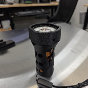 Könnte beinhalten: Eine schwarze Taschenlampe mit strukturiertem Griff und Lanyard. Die Taschenlampe hat kreisförmige Aussparungen und ein helles Licht an der Oberseite. Das Innere der Taschenlampe ist durch die Aussparungen sichtbar.