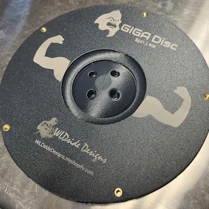 Pode incluir: Um disco circular preto Giga Disc com superfície texturizada, apresentando um cubo central com quatro orifícios, dois gráficos de braços flexionados e rebites metálicos dourados. O texto diz GIGA DISC, Spin 2 Win, WLDside Designs e WLDsideDesigns.myshopify.com.