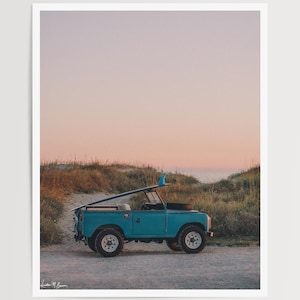 May include: A vintage teal-colored Land Rover with a surfboard mounted on top, parked on a sandy road near the beach. The vehicle has white wheels and is set against a backdrop of dunes and a pastel sky.