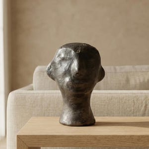 May include: A dark bronze-colored sculpture of a human head. The head has a rough, textured surface and is set on a light-colored wooden table. The sculpture is a decorative piece.
